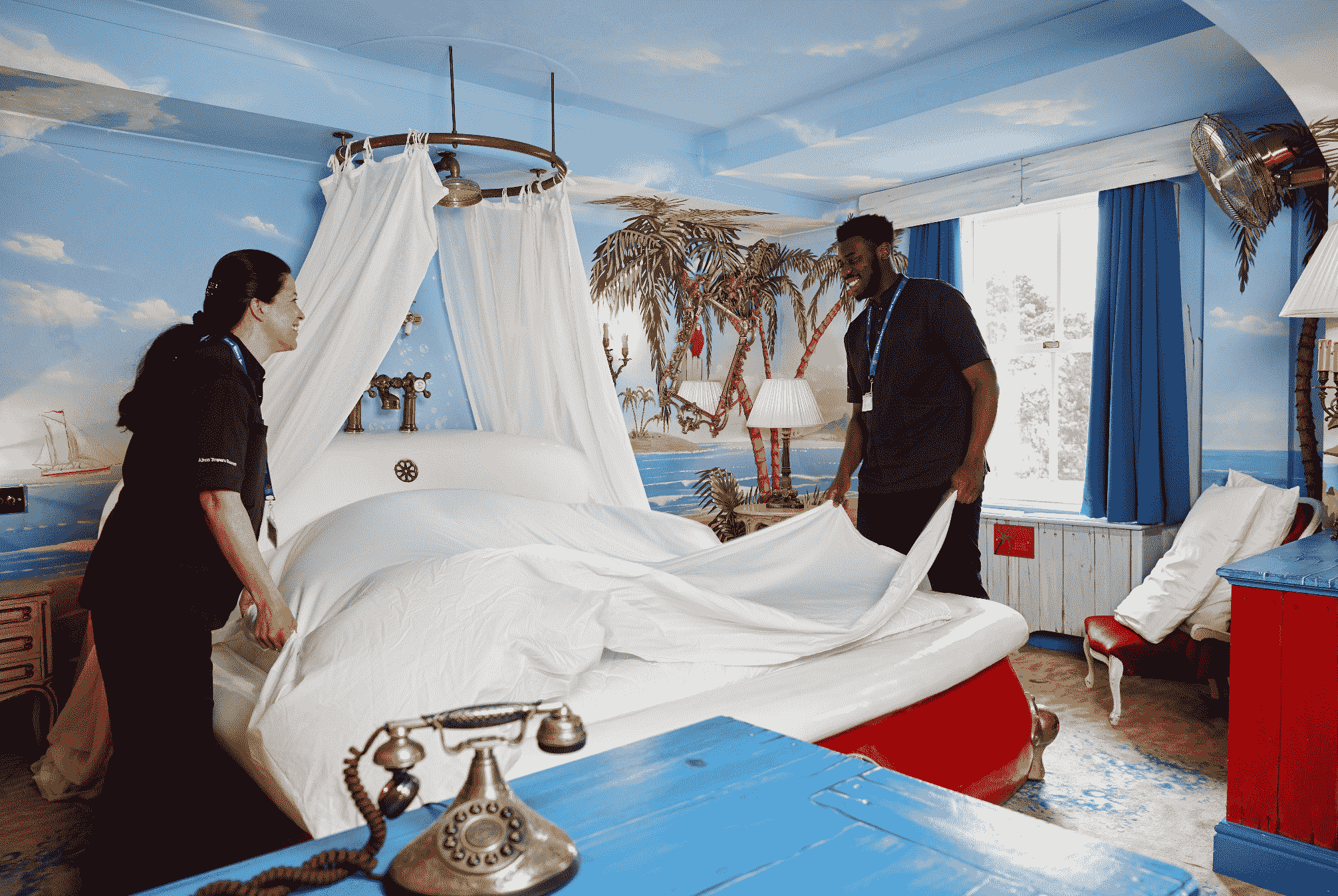 Two housekeepers are changing the white sheets of a bed in a sea themed hotel room.