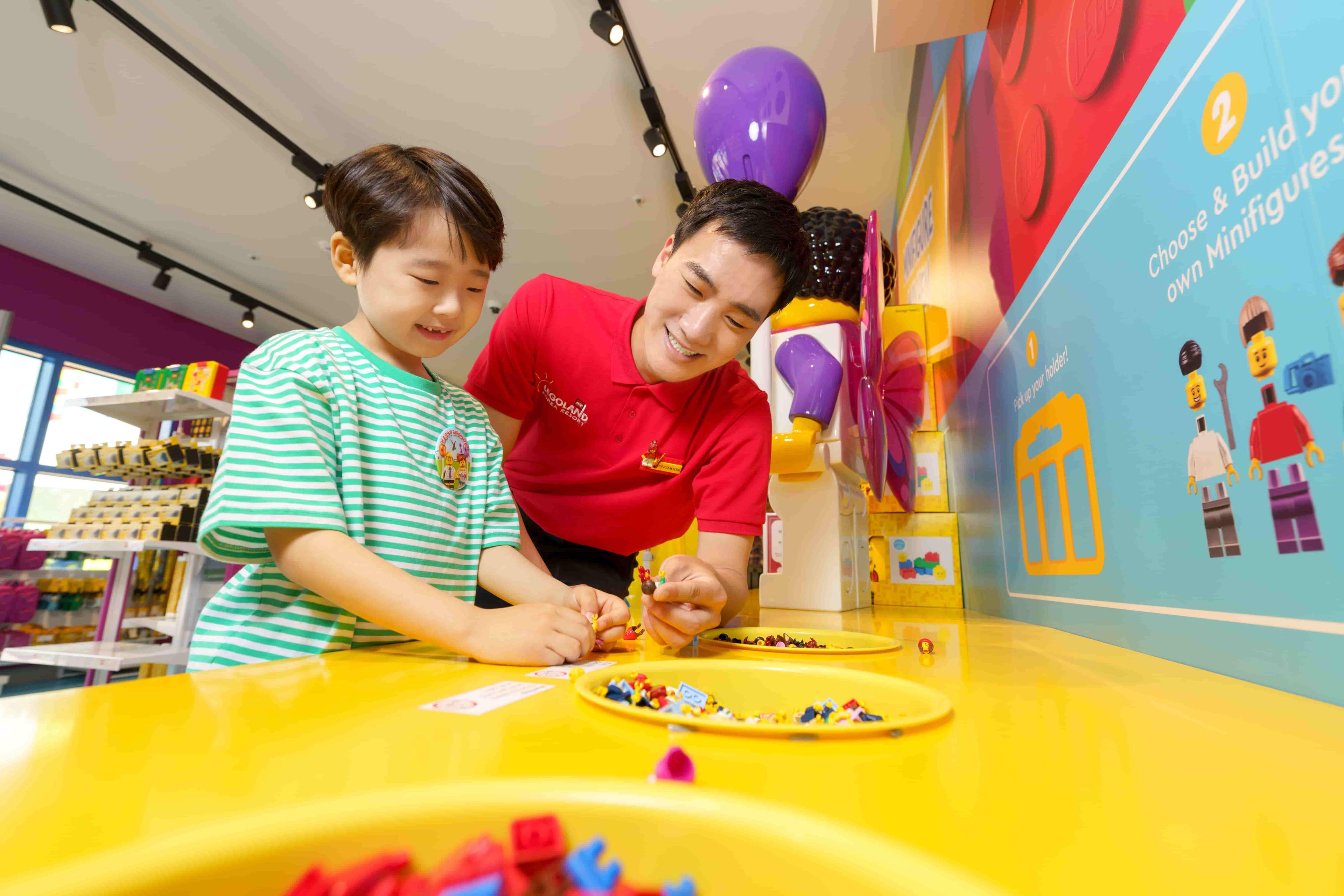 Man wearing a red polo shirt helps a young boy create his own LEGO® figure on a yellow table.