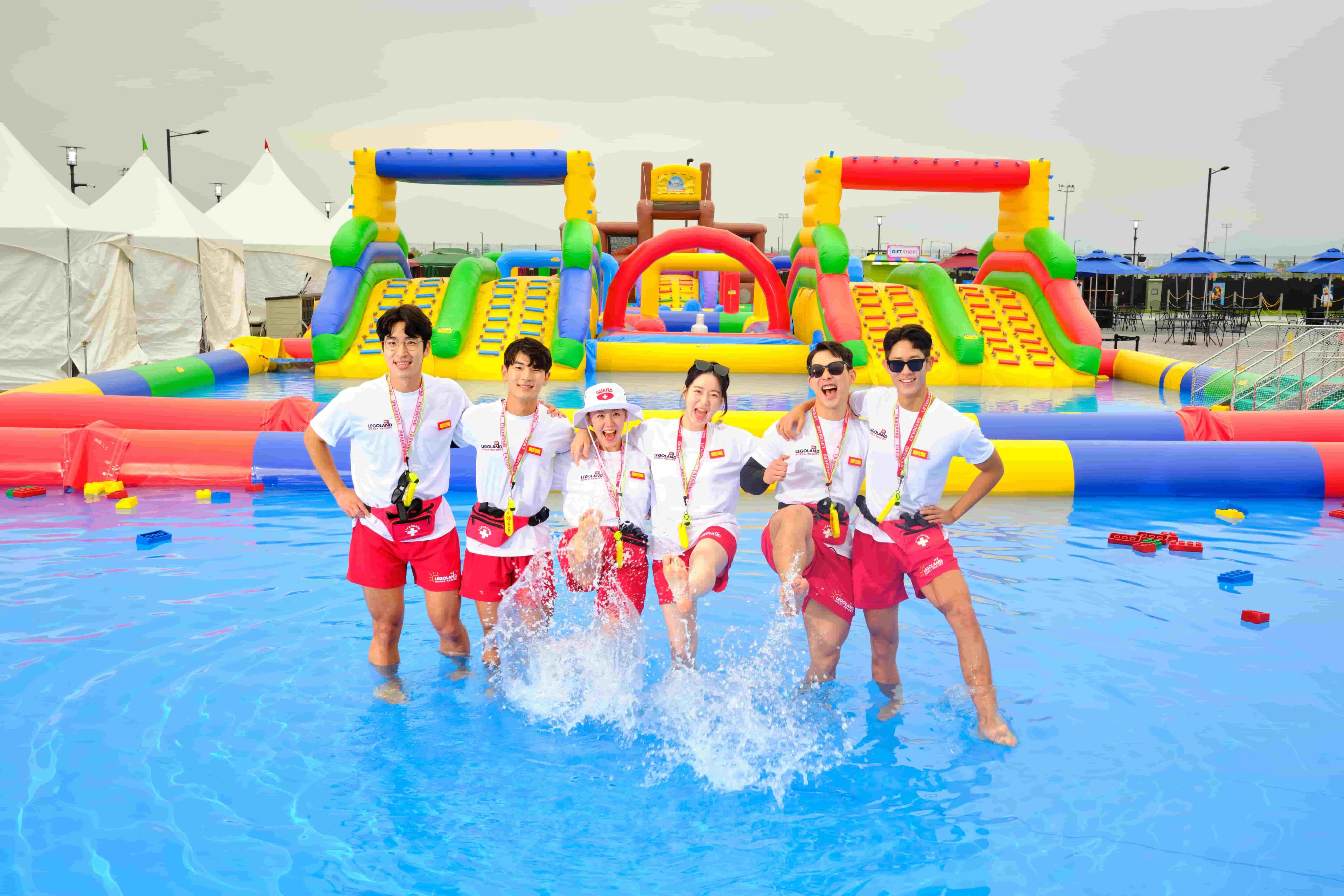 Group of lifeguards stand in the water kicking their feet towards the camera splashing. Behind them there is a bright inflatable obstacle course.