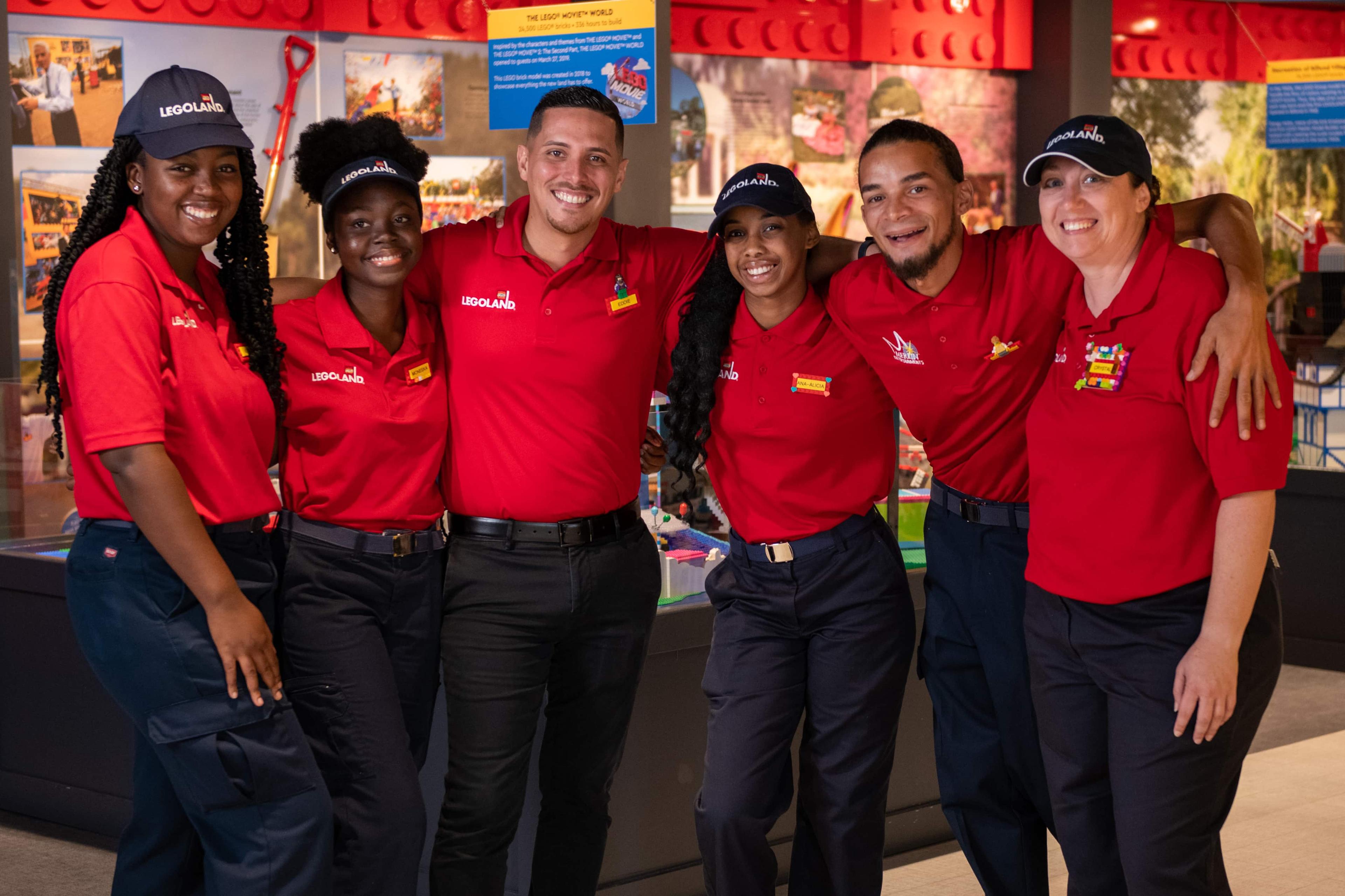 Five employees wearing red polo shirts stood in a line with their arms over each other. They are smiling and wearing black branded LEGOLAND® caps.