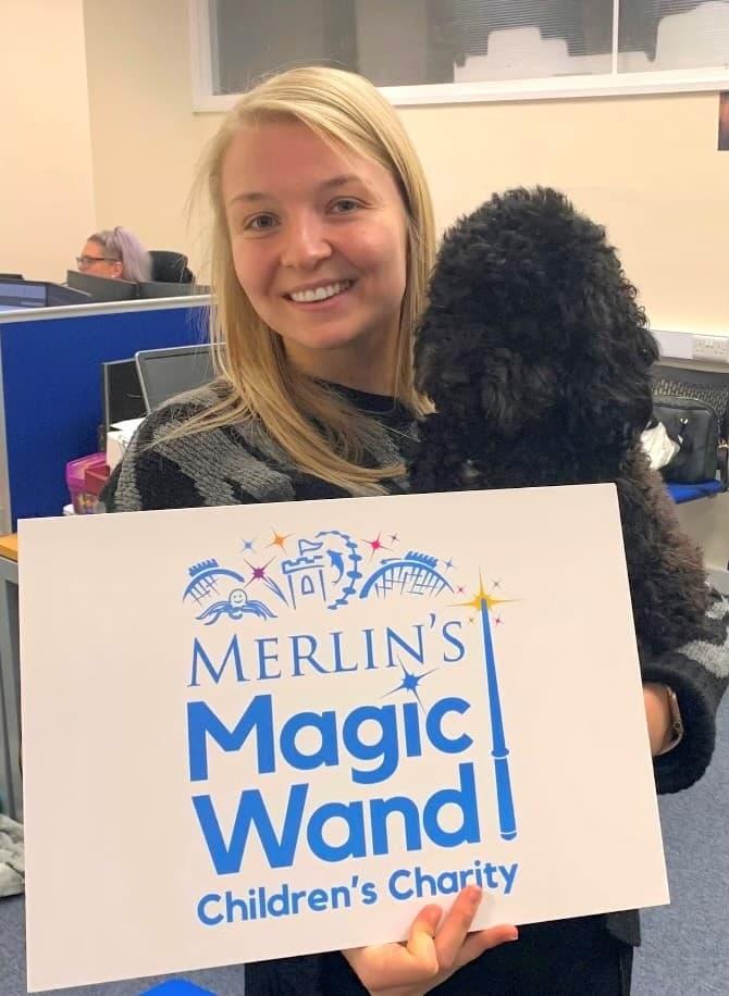 Blonde women holding a white sign with the text Merlin's Magic Wand Children's Charity in front of her. She is also holding a black fluffy dog.