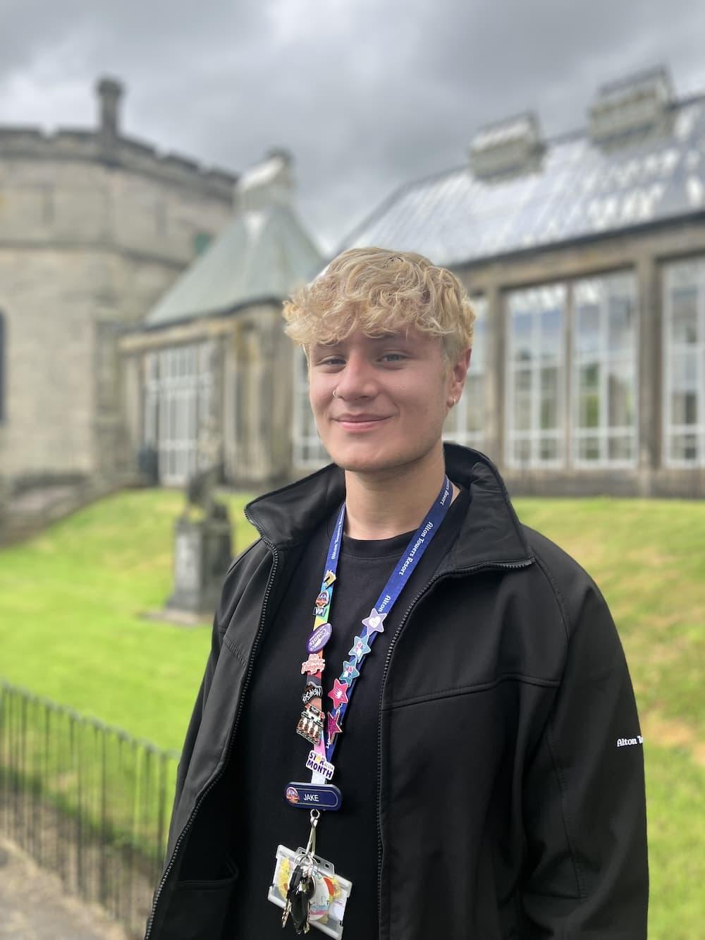 Blonde boy wearing a brown tshirt and jacket with a blue lanyard covered in badges stood in front