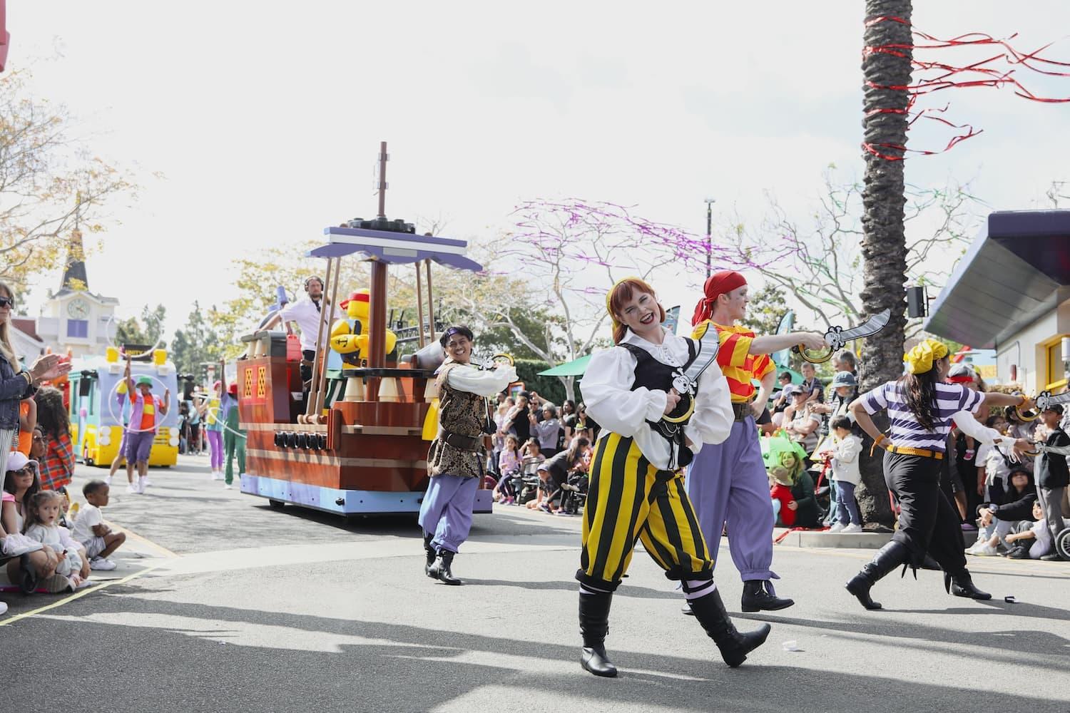 Actor in a pirate outfit is walking along a concrete road in a parade. Pirate ship float is in the background surrounded by actors and crowds.