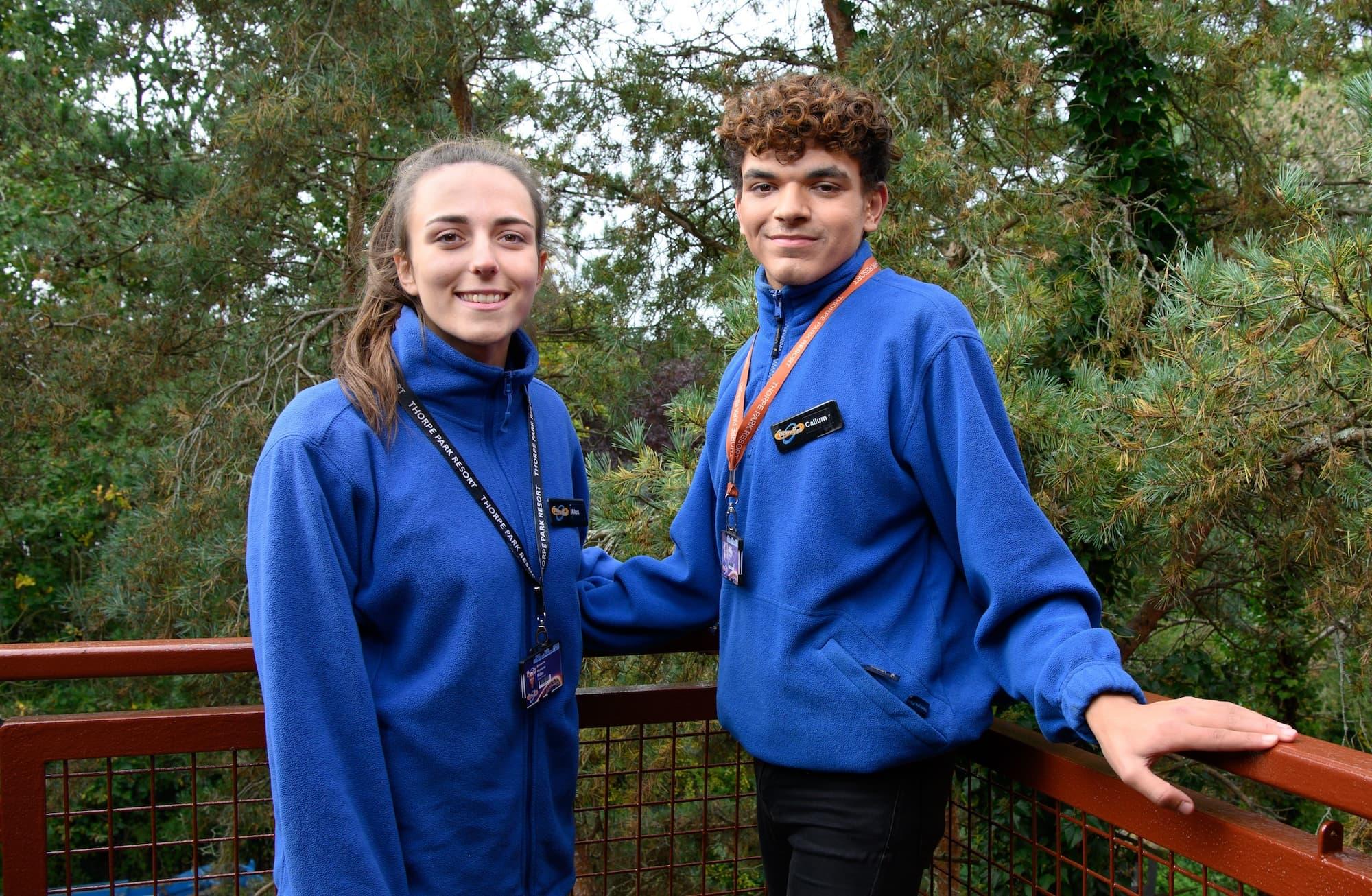 Man and women wearing blue Thorpe Park uniforms and lanyards are stood in front of a metal brown fence.