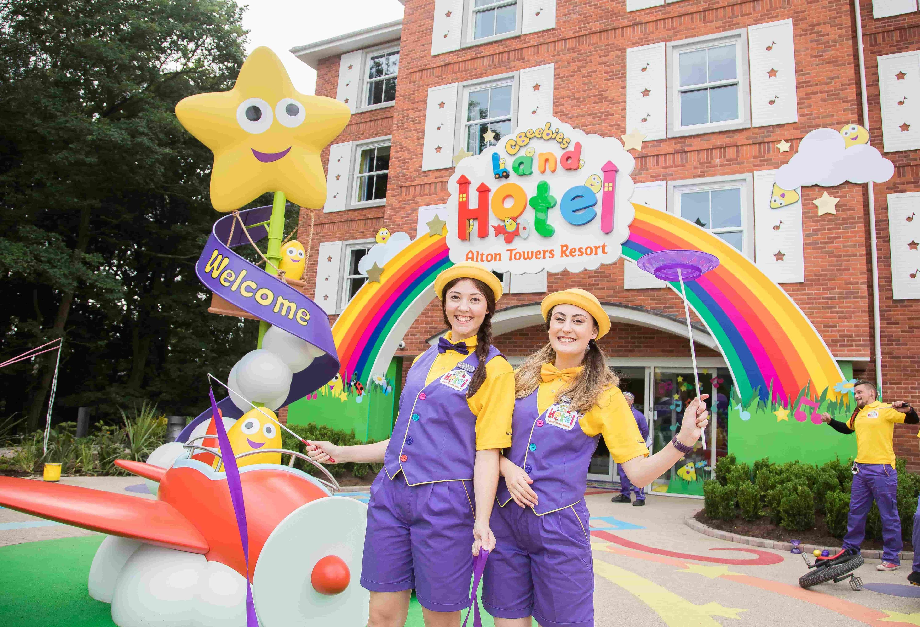 Two females wearing yellow polo shirts and bow ties and purple waistcoats and shorts and stood outside on the CBeebies Hotel building with a rainbow sign.