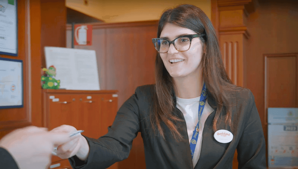 Women with brown hair and glasses is passing a hotel key card over a reception desk. She is wearing a white tshirt with a black blazer and a blue Gardaland lanyard.