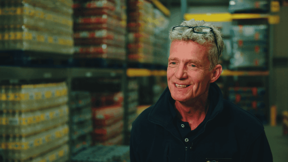 Man with grey hair and glasses is wearing a blue polo shirt stood in front of crates of stacked items in a warehouse.