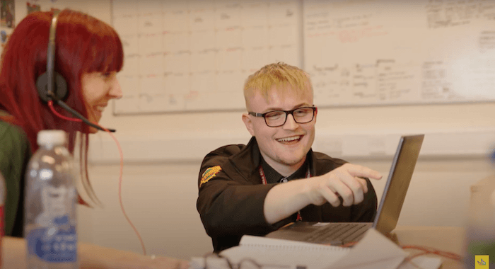 Blonde young man wearing glasses is pointing at a laptop screen. Women with red hair and a headset is sat next to him looking at the screen and smiling.