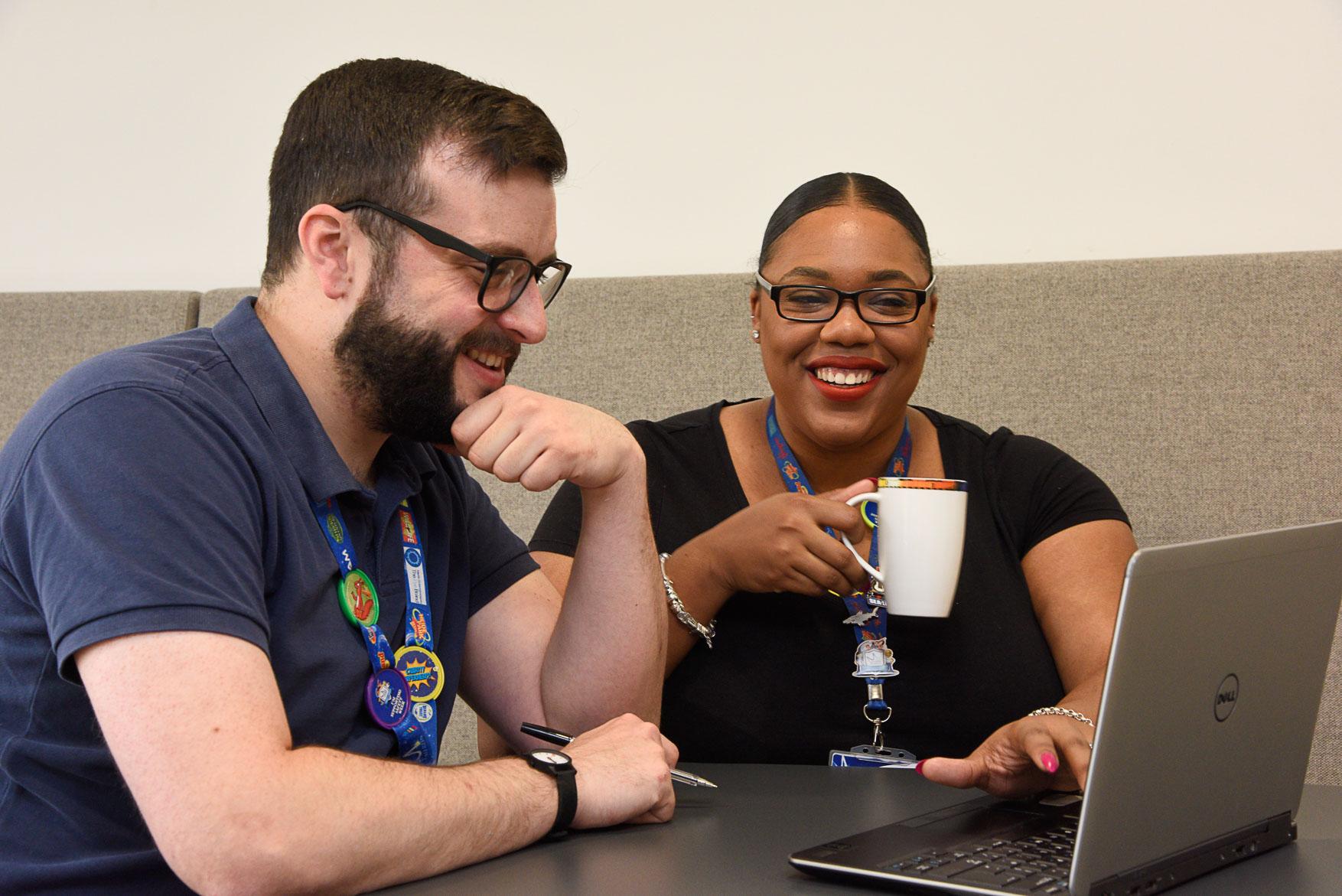 Two people sitting at a table smiling, one holding a mug and the other using a laptop, both wearing lanyards.