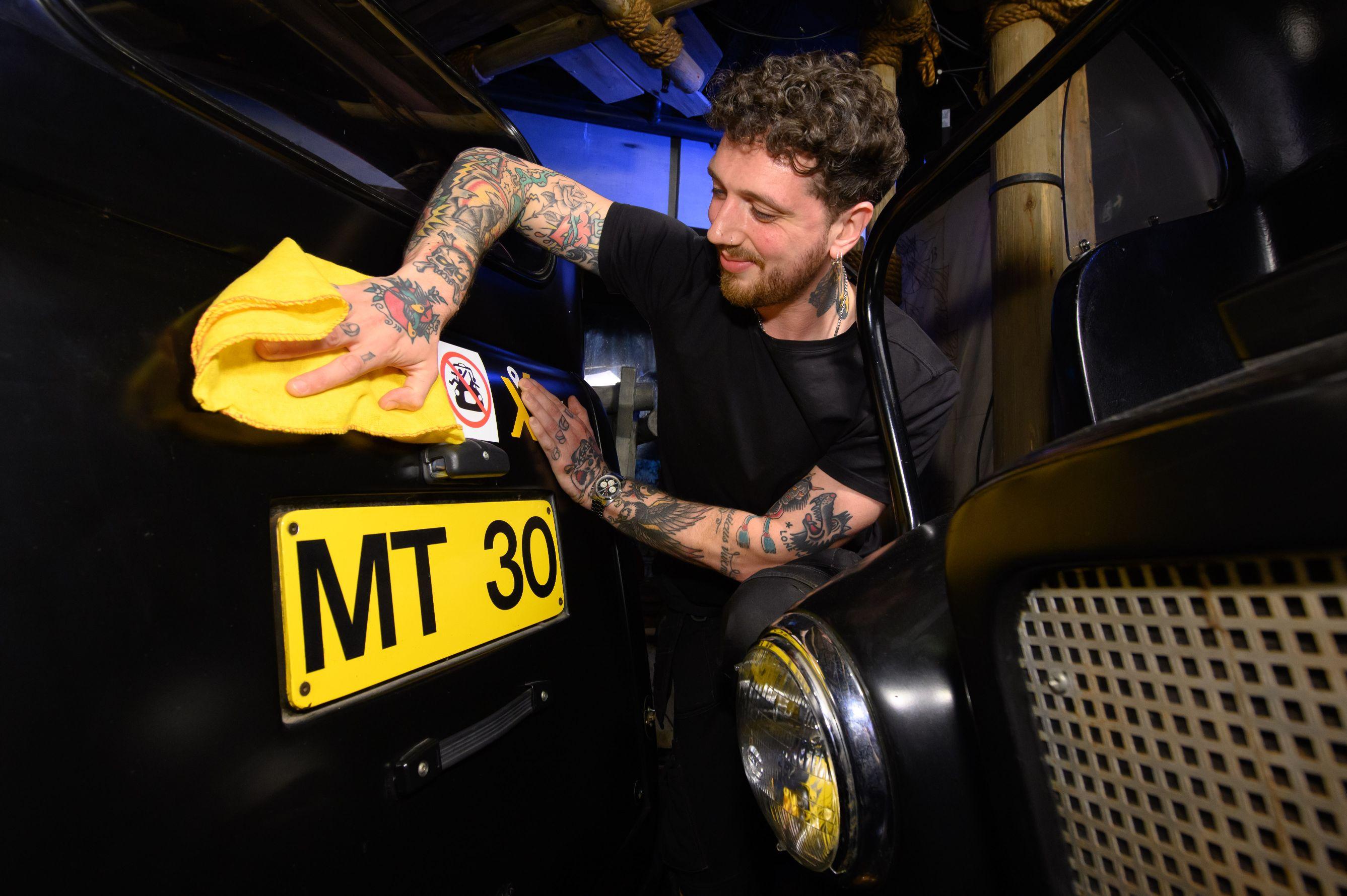 Person cleaning the front of a black vehicle with a yellow cloth, focusing on a yellow license plate that reads ‘MT 30’ and a badge above it.