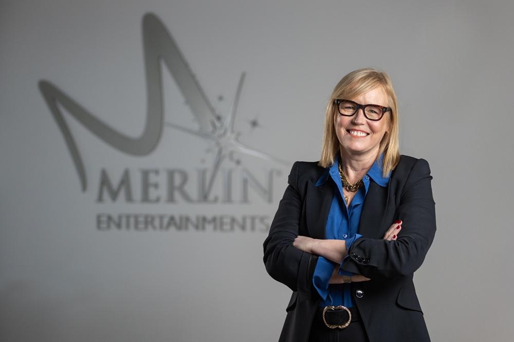 CEO of Merlin Entertainments, Fiona Eastwood, in a business suit standing with arms crossed, smiling in front of a wall displaying the Merlin Entertainments logo.