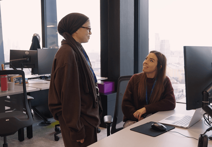 Two colleagues talking at a desk. One is stood and one is seated. They are both smiling. London skyline is visible behind.