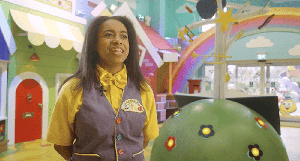 Women in a yellow and purple outfit smiling on a CBeebies set surrounded by colours and rainbows.